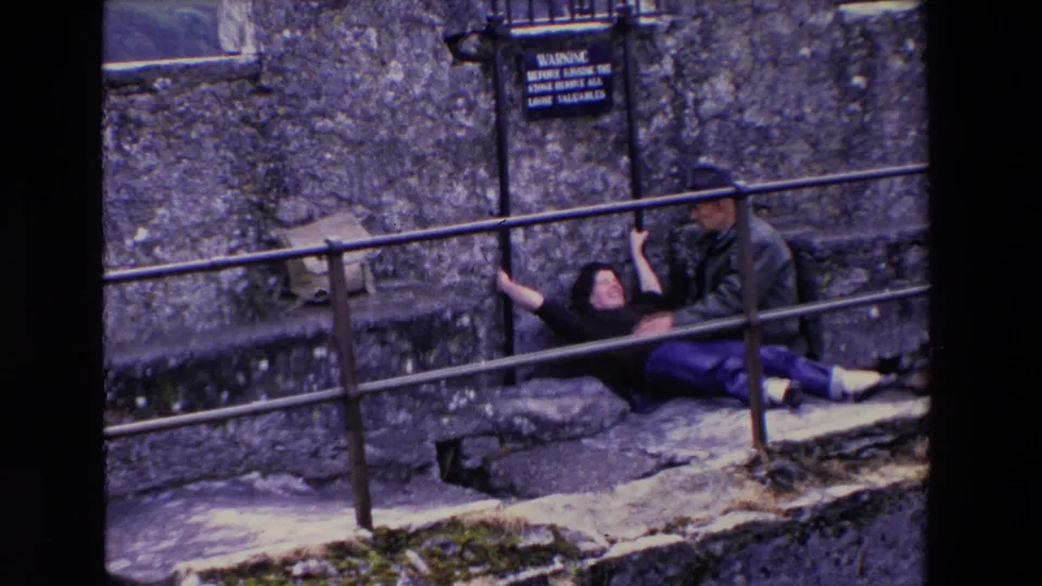In 1969 Ireland, a woman lies back near the Blarney Stone area while a man assists her, with castle stonework, a warning sign, and metal railings in view.