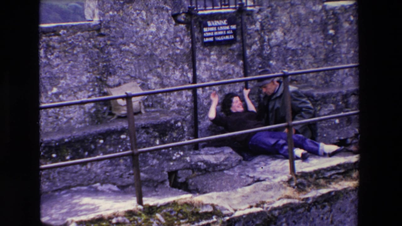 In a 1969 Ireland home-movie frame, a woman in dark clothing and purple pants playfully leans back from a metal railing while a man in a cap sits beside her against a stone wall.