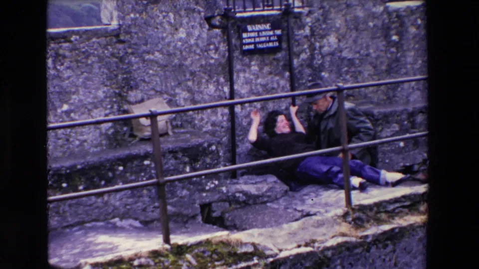 In a 1969 Ireland home-movie frame, a woman in dark clothing and purple pants playfully leans back from a metal railing while a man in a cap sits beside her against a stone wall.