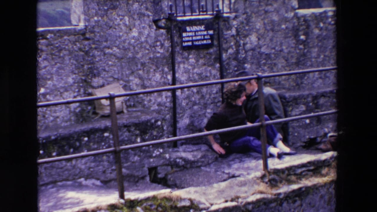 The woman and man sit close together on the stone steps in Ireland, with the woman turned toward him near the metal railing and a black warning sign on the wall behind them.