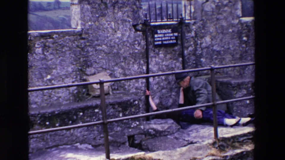 The woman appears partly reclined beneath the railing with her arms raised toward it, while the man remains seated beside her on the stone steps in the 1969 Ireland home-movie scene.