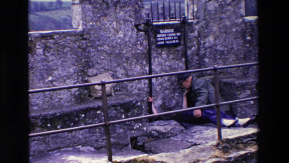 At the end of the clip, the man sits on the stone steps beside the partly hidden woman, with the metal railing crossing the foreground and the warning sign still visible on the wall.