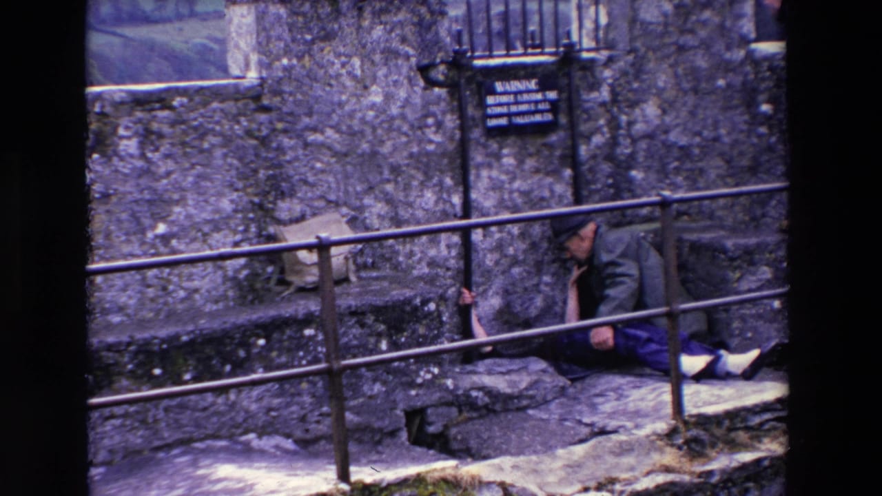 In supplied 1969 Ireland 8mm footage, an older man in a cap sits on stone steps near a metal railing, rough stone wall, barred window, and warning sign.
