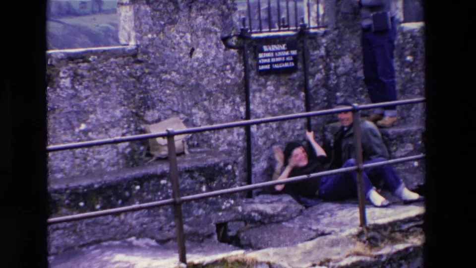 In supplied 1969 Ireland home-movie footage, an older woman lies back laughing or gesturing on stone steps while an older man sits nearby and another visitor's legs appear above them.