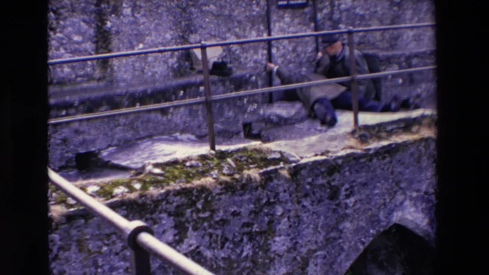 In supplied 1969 Ireland 8mm footage, the older couple remains beside the metal railing on stone steps, with the man seated and the woman reclining near the rough wall and warning sign.