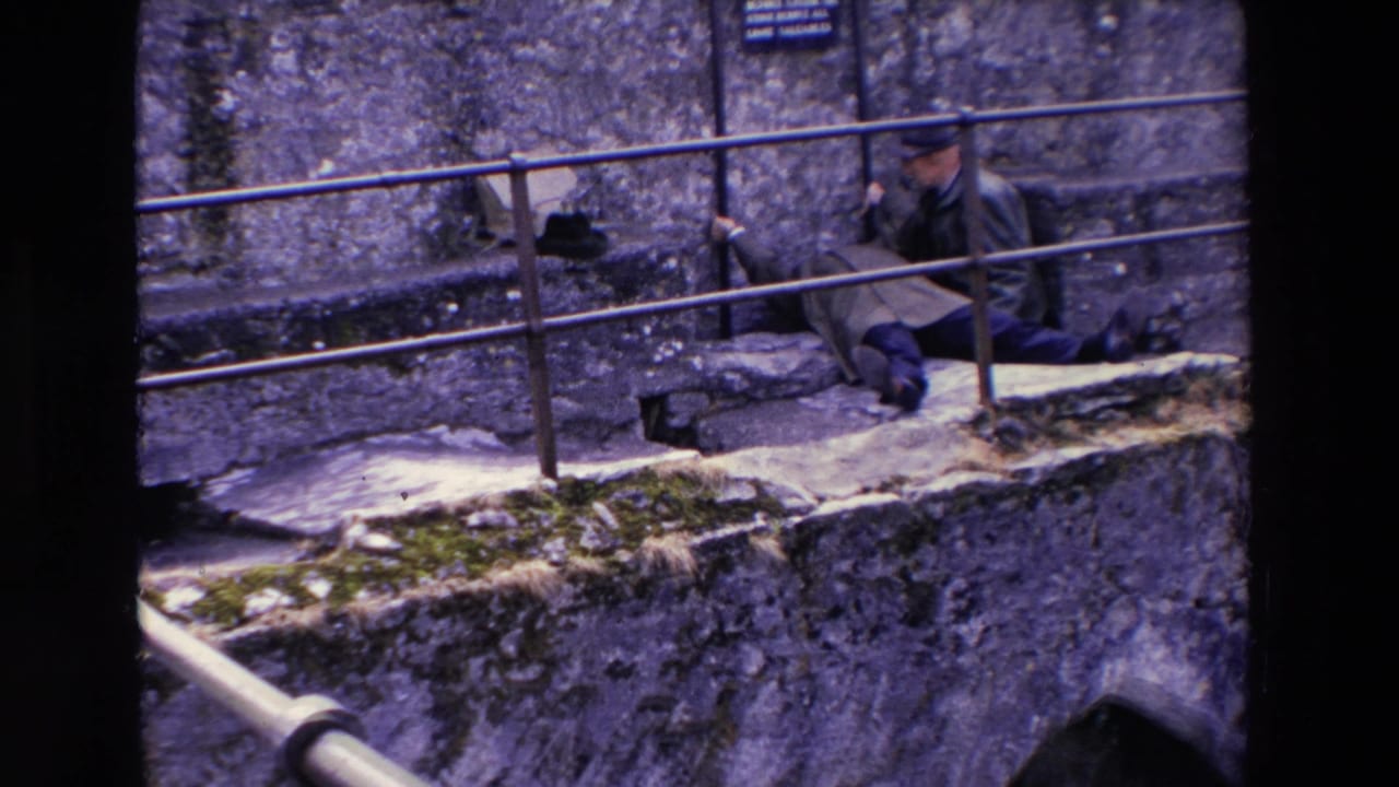 In 1969 Ireland home-movie footage, two adult men are on a mossy stone ledge beside a metal railing and rough stone wall, with one man seated low and another nearby.