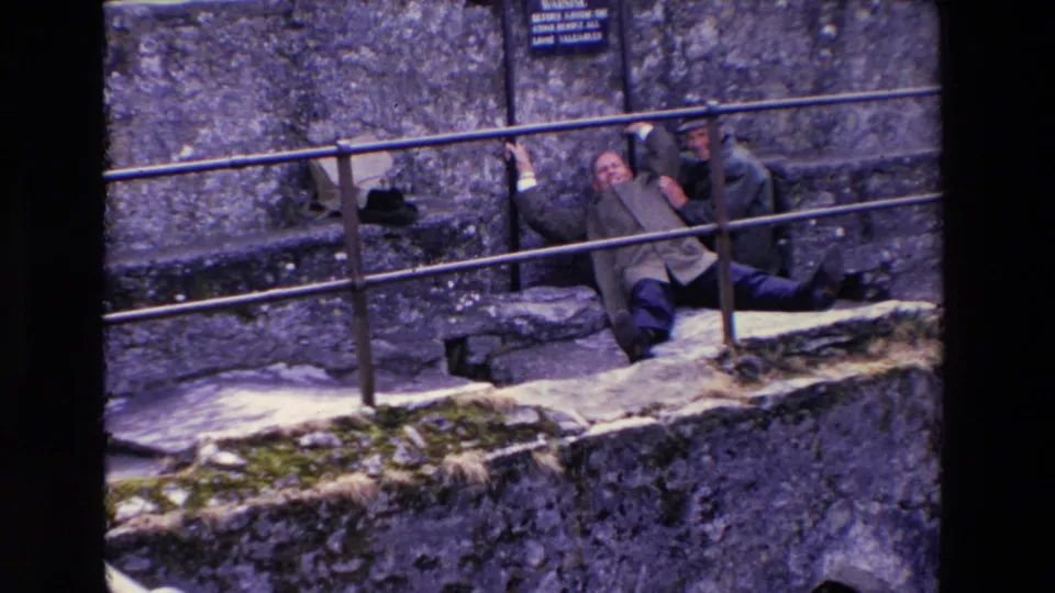 The man on the ledge raises an arm while sitting against the stonework, with his companion close by and a warning sign visible above the railing.