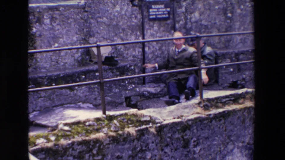 By the end of the brief 1969 Ireland clip, the man appears seated upright on the stone ledge near the railing, with another adult man partly visible behind him.