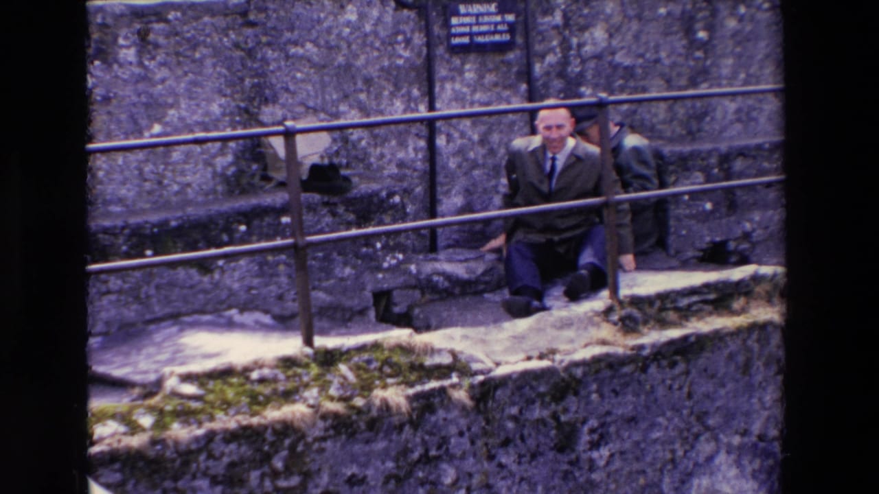 In 1969 Ireland home-movie footage, a suited man sits on stone steps near a metal railing and weathered masonry, with another adult partly visible behind him.