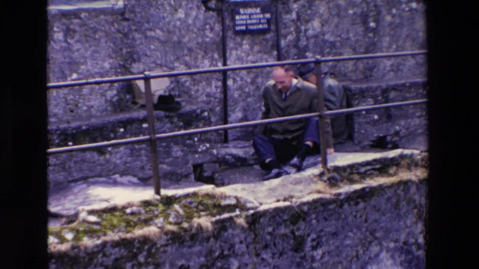 A suited adult man appears to sit or scoot along stone steps by a railing at an Irish castle ruin, with rough walls and a small warning sign behind him.