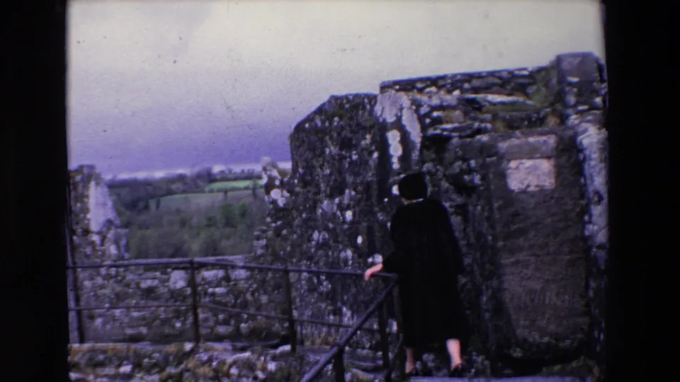 A woman in a dark coat stands beside a metal railing at a castle ruin in Ireland, facing outward toward green fields beyond the stone walls.