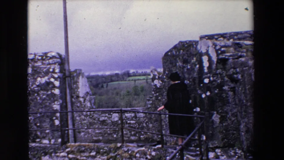The home-movie frame shows a woman at the edge of an Irish castle ruin, holding or standing near the railing while looking toward the rural landscape.