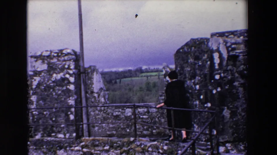 A woman in a dark coat stands with her back partly turned on castle ramparts in Ireland, with rough stone walls and green countryside beyond.