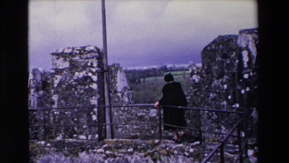 The woman remains on the castle walkway in Ireland, framed by weathered stone walls, a metal railing, and distant fields under a cloudy sky.