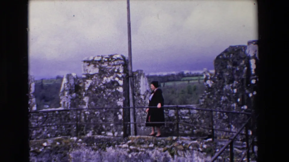 A woman in a black coat over a red dress walks along the castle ramparts in Ireland, with stonework, railing, and rural landscape visible around her.