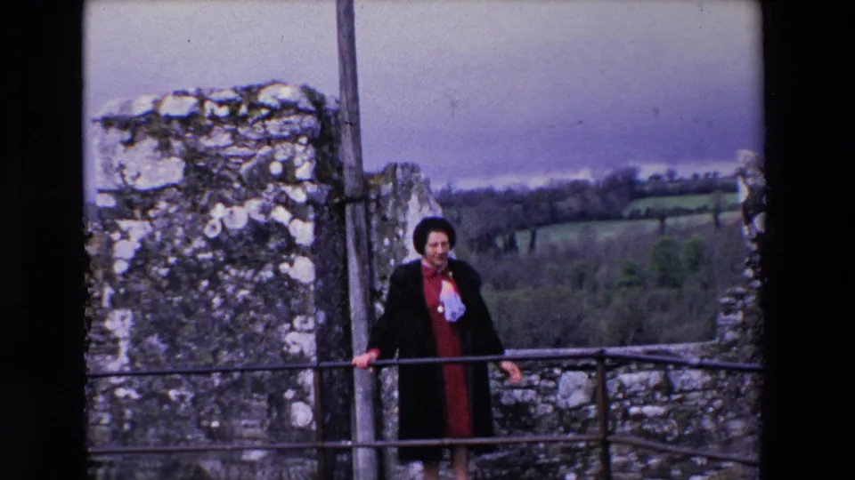 The woman faces the camera on the castle ramparts in Ireland, holding the metal railing with rough stone walls and green countryside behind her.