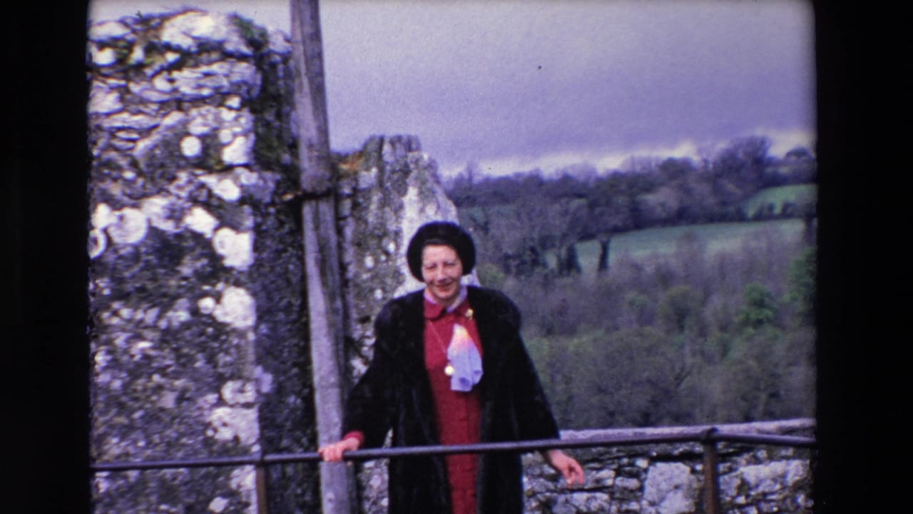 In 1969 Ireland, a woman wearing a dark coat, dark hat, red dress, and light scarf stands on a stone balcony beside old walls with a rural landscape in the background.