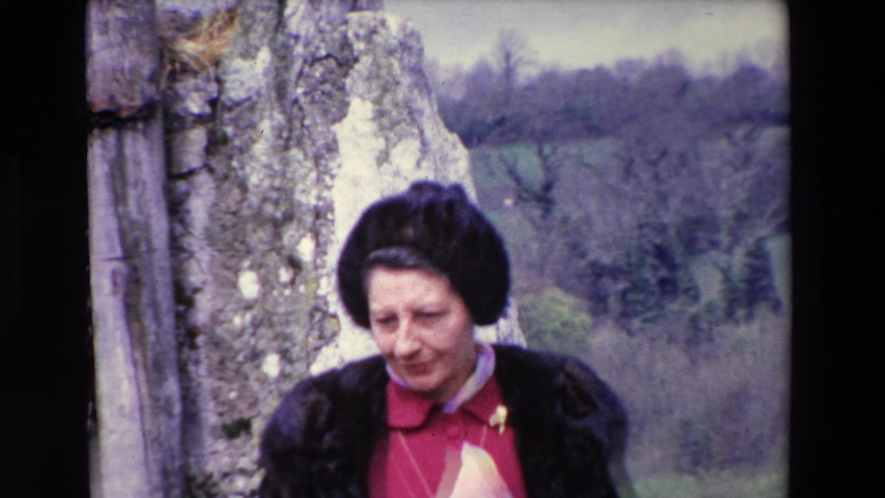 In 1969 Ireland, a closer home-movie frame shows the woman near a weathered stone wall, with her dark hat, red dress, light scarf, and muted countryside visible behind her.