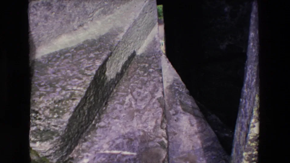 A handheld 1969 Ireland frame looks downward along stone paving and rough ruin walls, with only part of the nearby figure visible at the right edge.
