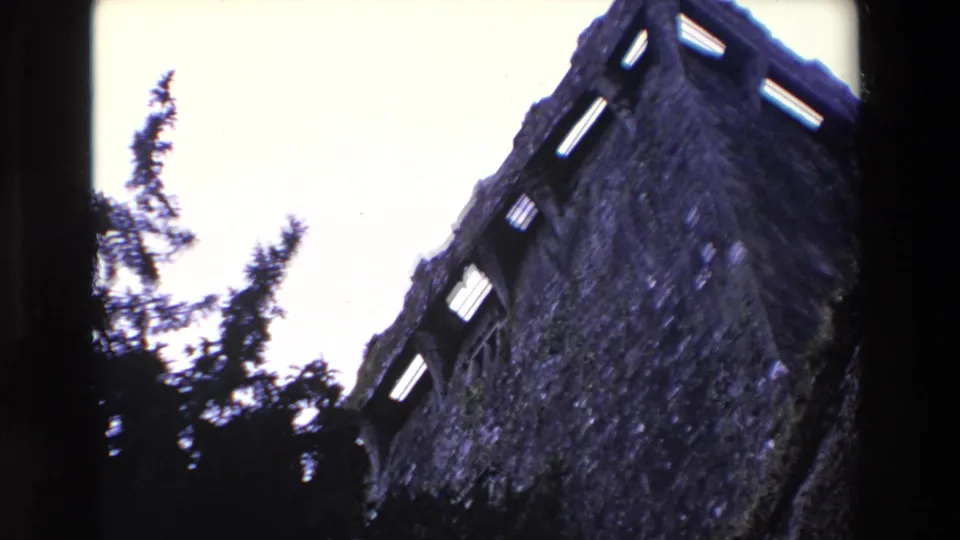 1969 Ireland 8mm frame of a tall weathered stone tower or ruin wall, with open window slots, trees, and pale sky.