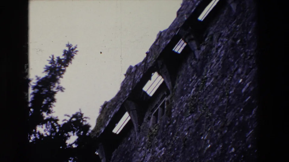1969 Ireland 8mm frame with the camera closer to the castle wall, showing rough masonry, bright arched openings, dark evergreens, and a vertical film-frame edge.
