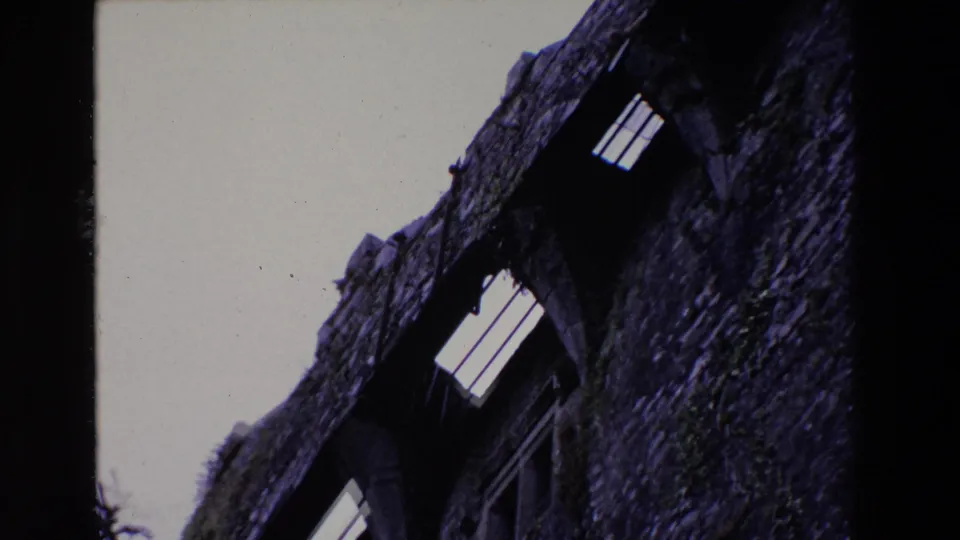 1969 Ireland home-movie frame looking up along a castle wall with textured stone, bright narrow openings, a pale sky, and heavy shadow from the handheld 8mm exposure.