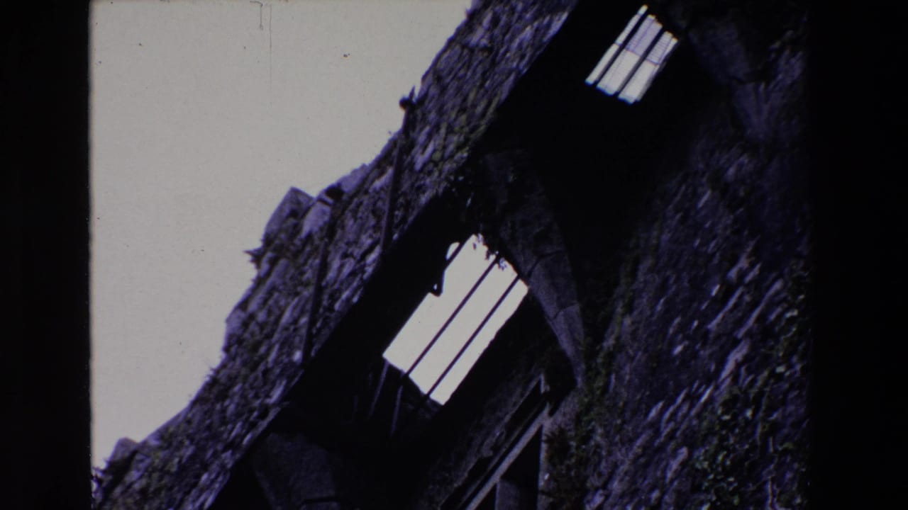 1969 Ireland 8mm frame showing a tilted low-angle view of rough grey masonry, dark arched openings, and a small barred opening against overcast sky.