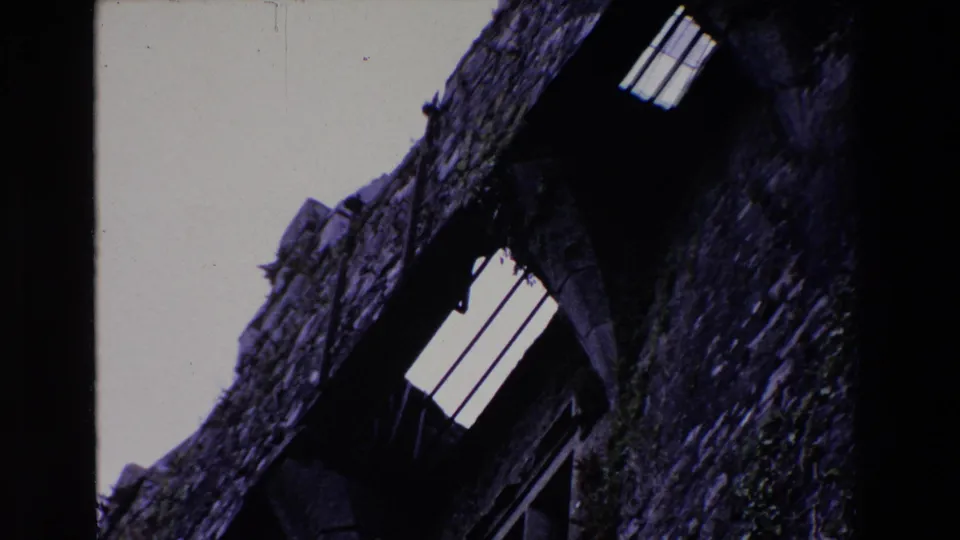 1969 Ireland 8mm frame showing a tilted low-angle view of rough grey masonry, dark arched openings, and a small barred opening against overcast sky.