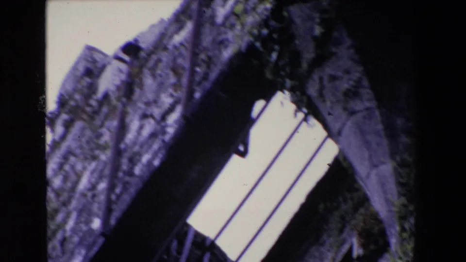 1969 Ireland 8mm frame showing a soft, tilted view upward through a barred opening in an old stone wall, with dark masonry and pale sky dominating the frame.