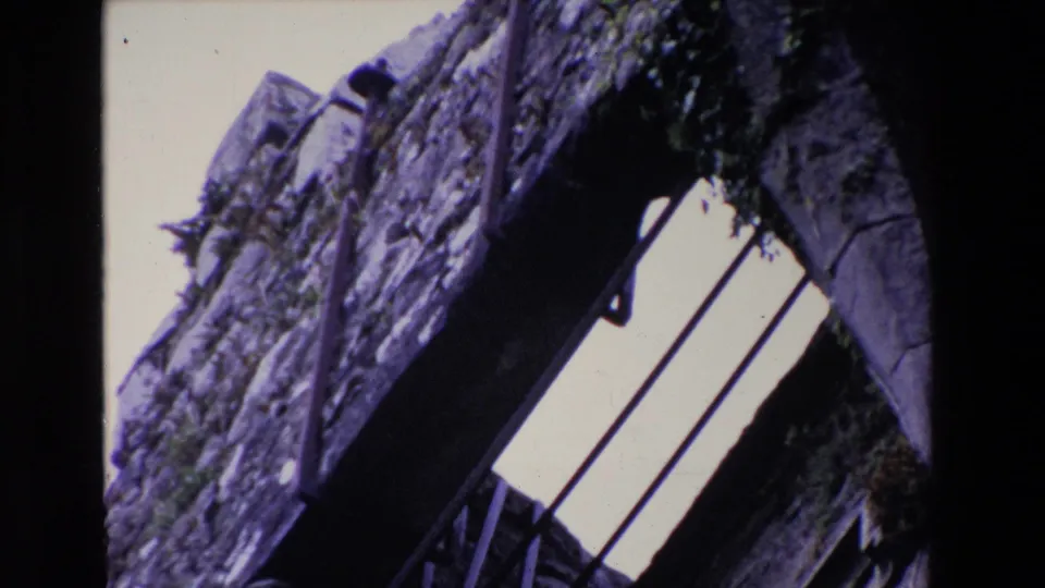 1969 Ireland 8mm frame showing the weathered upper portion of a stone structure, with a barred arched opening, rough masonry, greenery, and grey daylight.