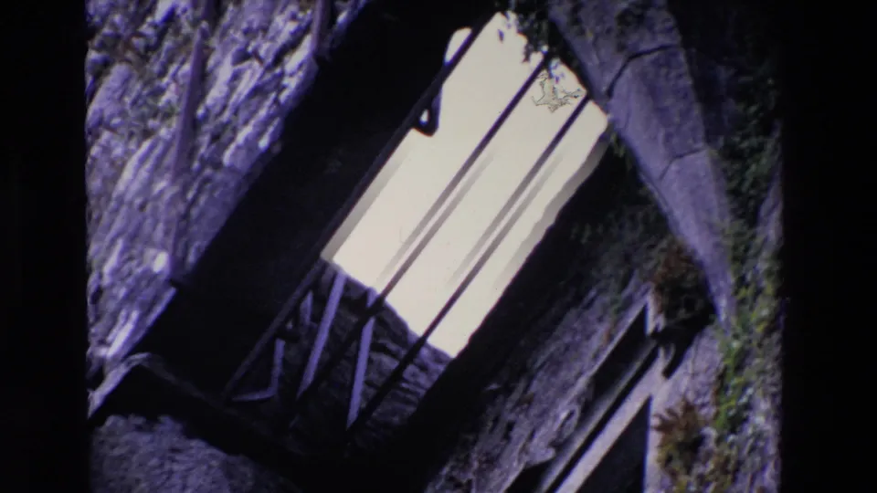1969 Ireland 8mm frame with rough masonry, a narrow barred window, surrounding shadow, and patches of vegetation on the wall.