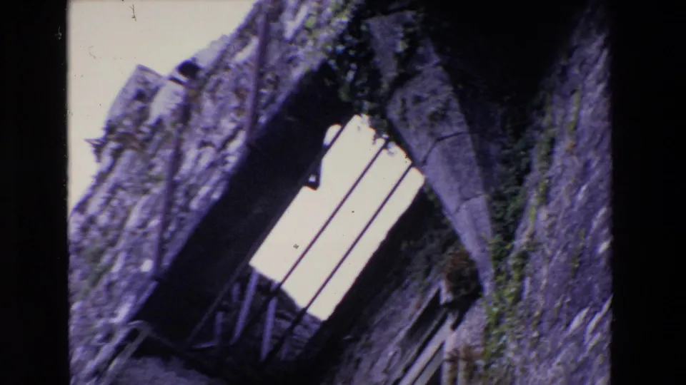 1969 Ireland 8mm frame showing the camera tilted along an old stone facade with a bright barred window, arched stonework, and greenery.