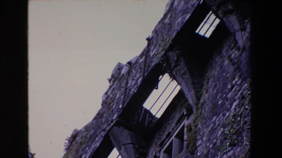 1969 Ireland 8mm frame showing a wider tilted view of the stone structure, including small barred windows, dark openings, and moss along the wall.