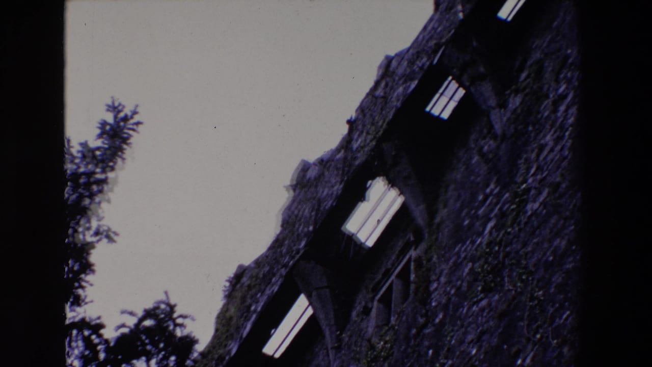 1969 Ireland 8mm frame showing a low-angle view of a mossy rough-stone building wall with dark arched window openings against a pale sky.
