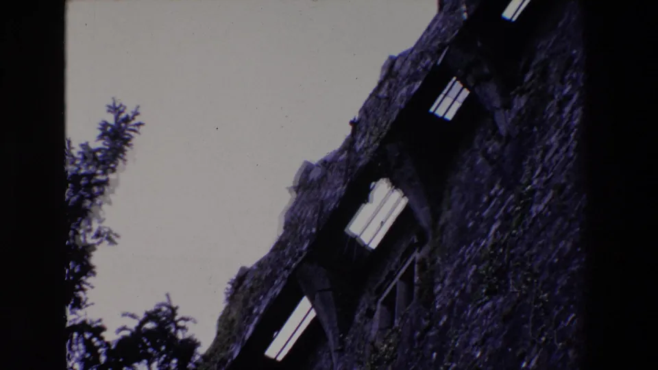 1969 Ireland 8mm frame showing a low-angle view of a mossy rough-stone building wall with dark arched window openings against a pale sky.