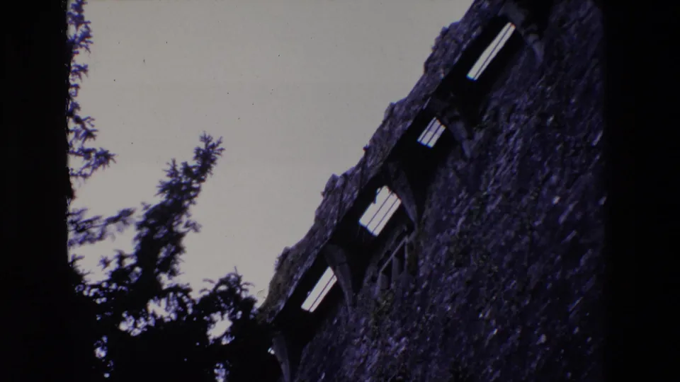 1969 Ireland 8mm frame with a tall weathered stone wall, bright arched window openings, and evergreen silhouettes rising in the foreground.