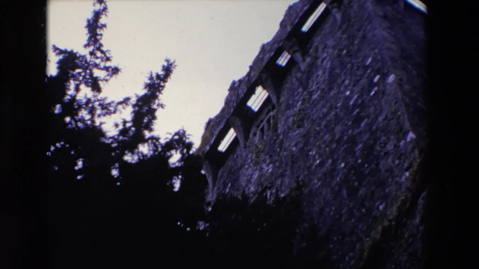 1969 Ireland 8mm frame showing the upper portion of a weathered stone building with arched openings, mossy wall texture, and dark evergreen silhouettes below.