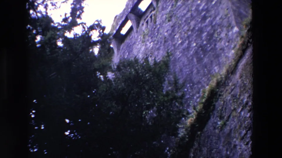 1969 Ireland 8mm frame with a tall rough stone wall, narrow arched window openings, and dense trees filling the lower view.