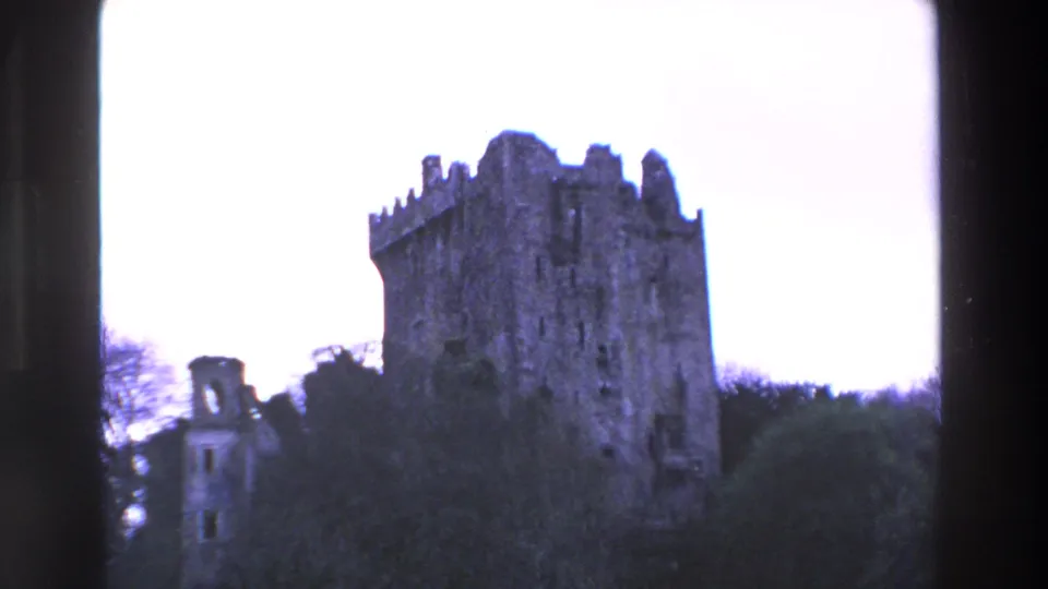1969 Ireland 8mm frame showing a crenellated stone castle tower with a smaller ruin nearby, partly obscured by trees and dark frame edges.