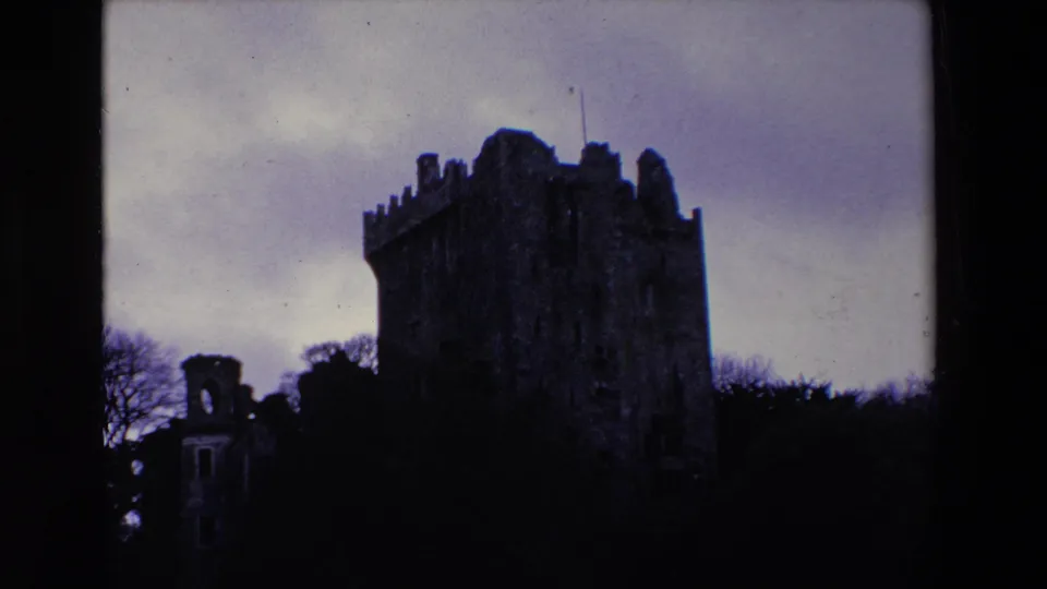 1969 Ireland 8mm frame showing the stone castle tower shifted left in frame, with a small ruined structure and dense trees below.