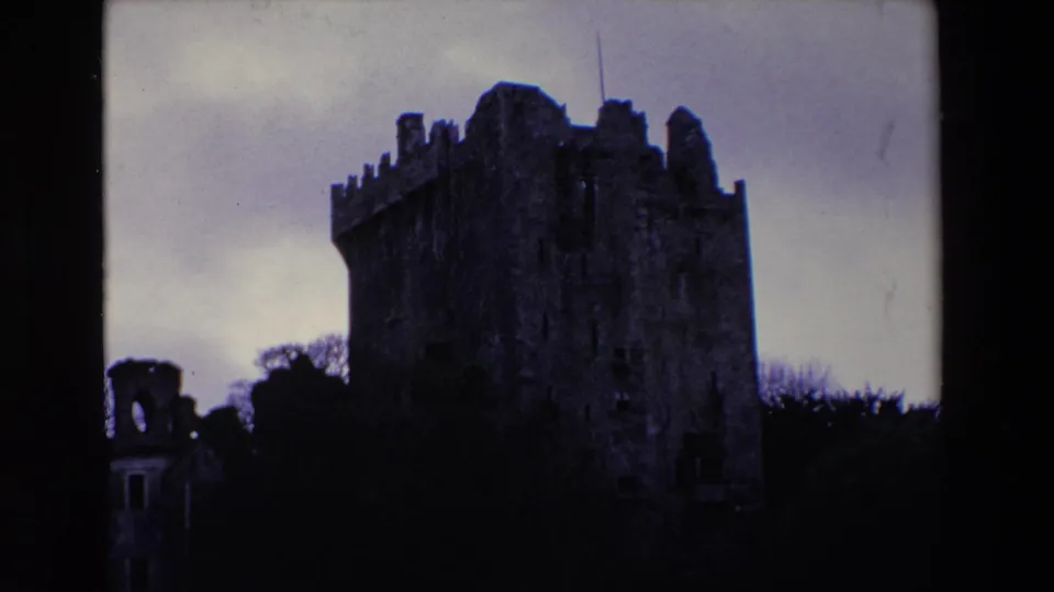 1969 Ireland 8mm frame showing the castle tower at right with battlements, a small ruin at left, dark trees, and grey cloud cover.