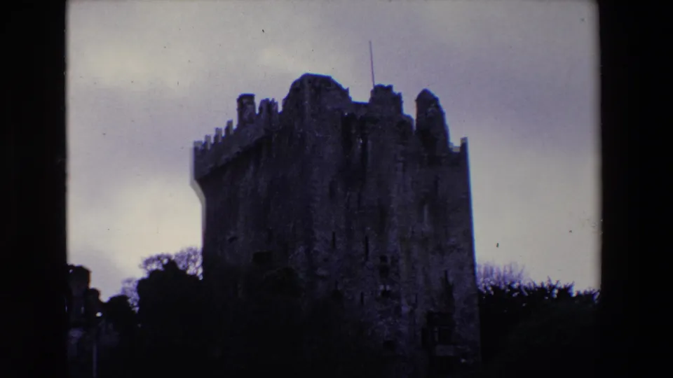 1969 Ireland 8mm frame showing a dark, weathered stone castle with crenellated towers silhouetted against a pale overcast sky.