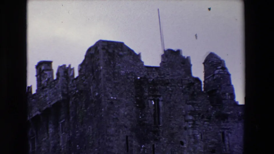1969 Ireland 8mm frame showing the rough stone castle wall, ruined battlements, narrow window openings, and a flagpole against a pale sky.
