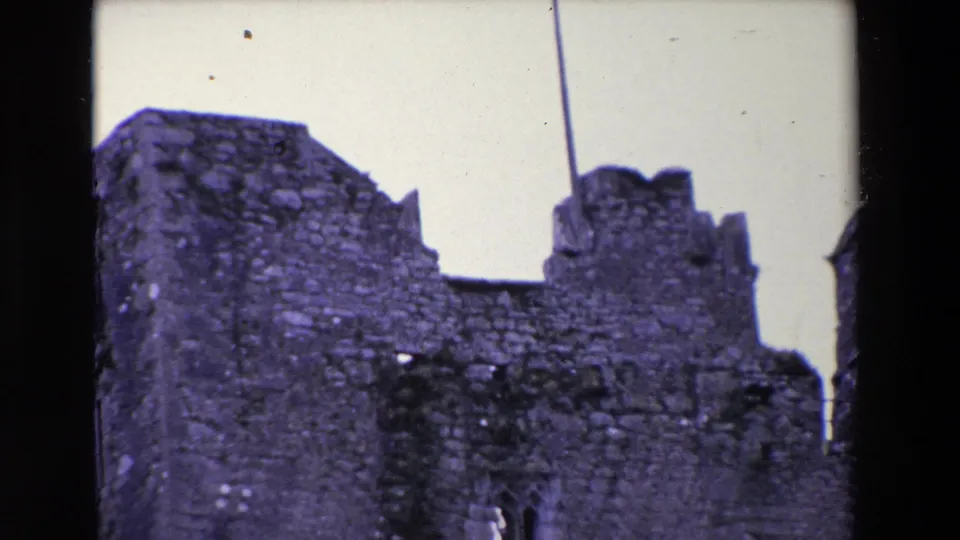1969 Ireland 8mm frame ending on a close view of weathered castle stonework, broken battlements, and a tall pole rising into the overcast sky.