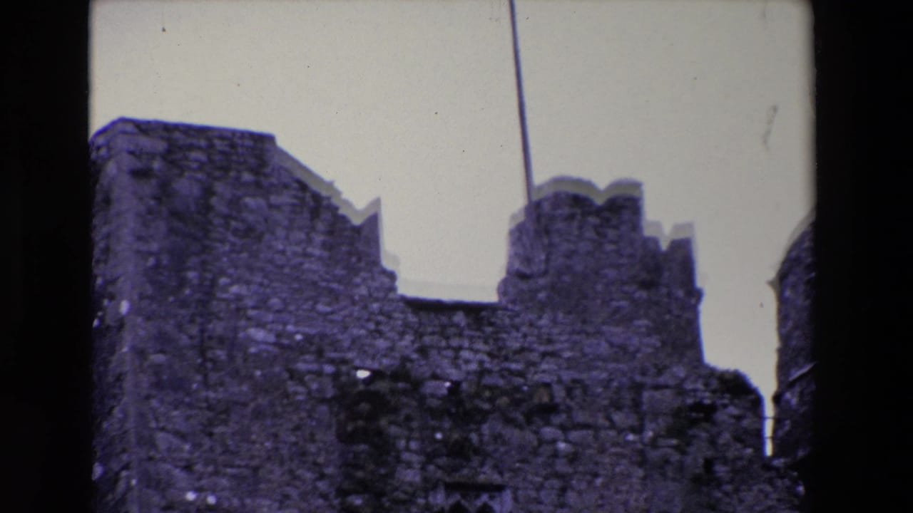 1969 Ireland home-movie frame showing a weathered stone ruin with crenellated walls and a vertical flagpole or mast line against a pale sky.