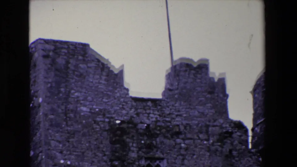 1969 Ireland home-movie frame showing a weathered stone ruin with crenellated walls and a vertical flagpole or mast line against a pale sky.