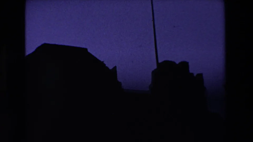 1969 Ireland home-movie frame where the castle ruin falls into shadow, creating a silhouetted outline below a blue-purple sky and visible film borders.