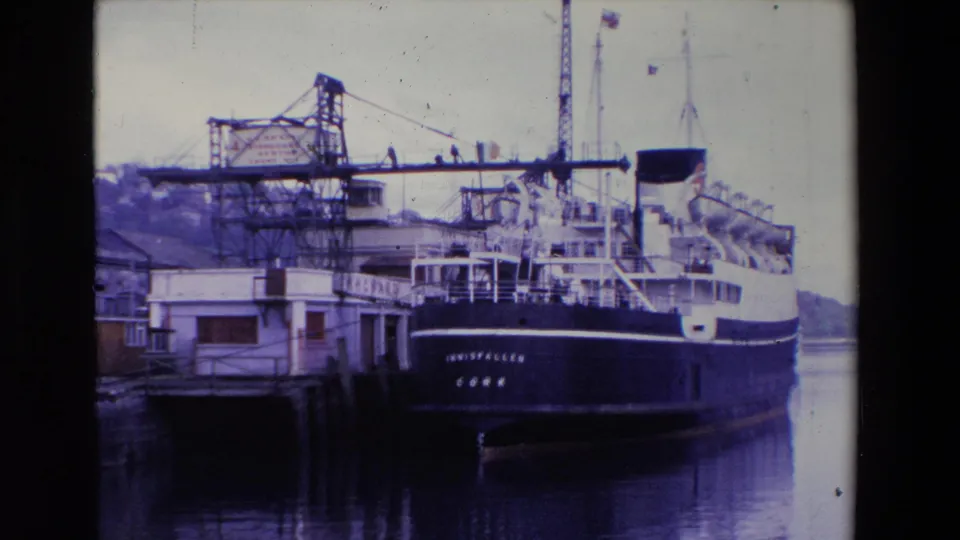 Wider 1969 Irish port view of the INNISFALLEN stern moored beside dock buildings, water, a crane, deck structures, and small distant figures.