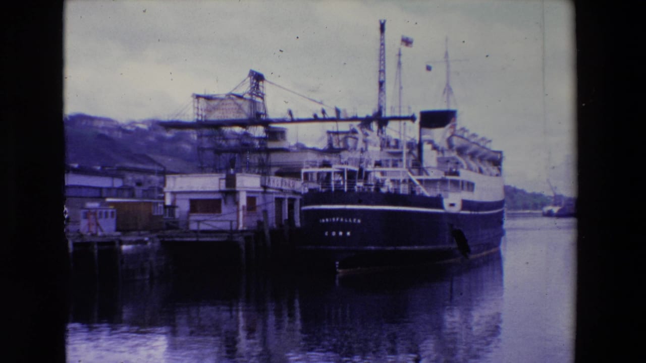 Ireland 1969 8mm frame showing the passenger ferry INISFALLEN moored at a wooden pier beneath an overcast sky, with cranes and hillside buildings behind it.