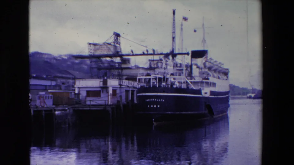 Ireland 1969 8mm frame showing the passenger ferry INISFALLEN moored at a wooden pier beneath an overcast sky, with cranes and hillside buildings behind it.
